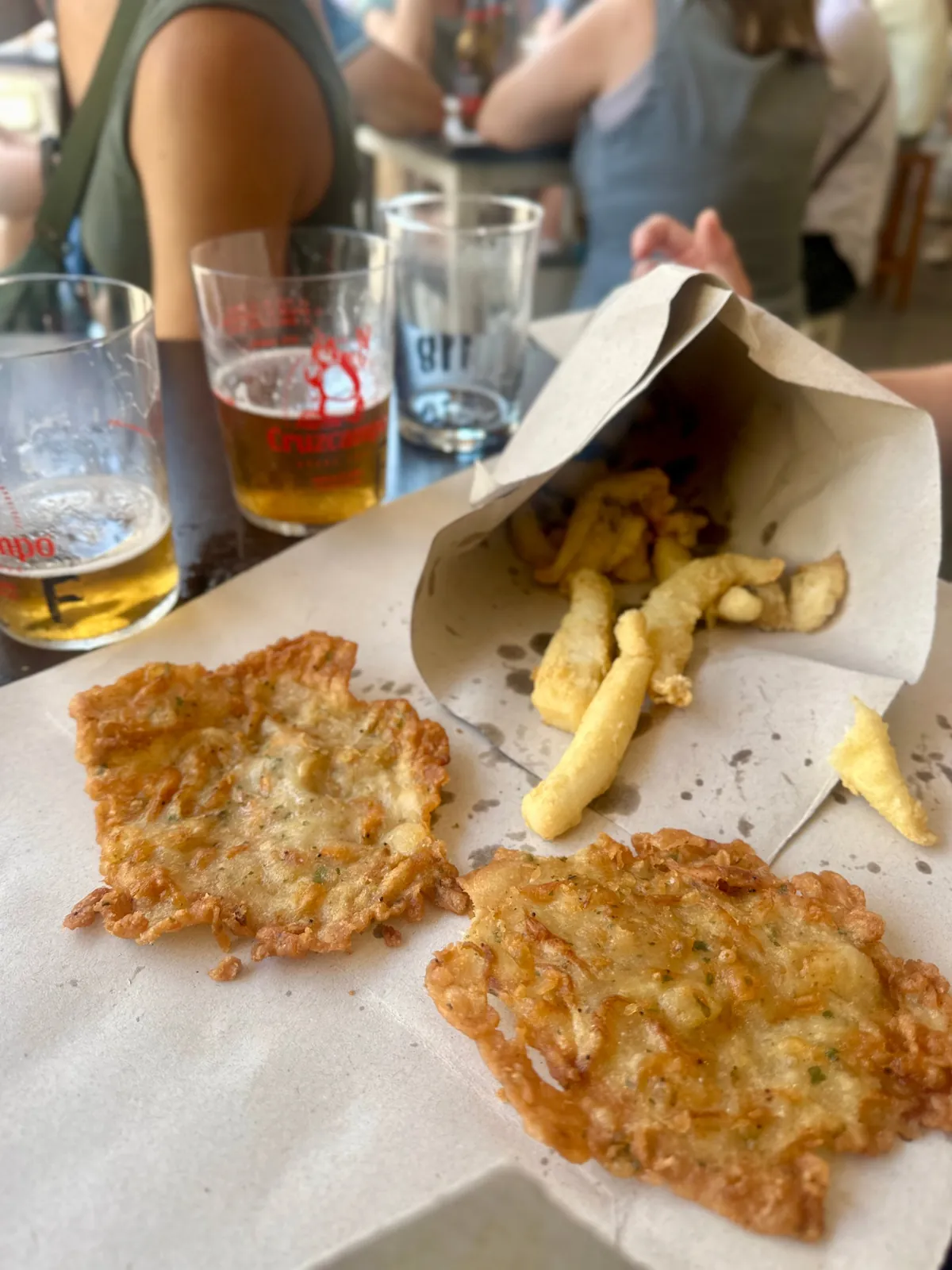 Tortillitas de camarones und frittierter Fisch auf Papier, daneben Bier in Plastikbechern.