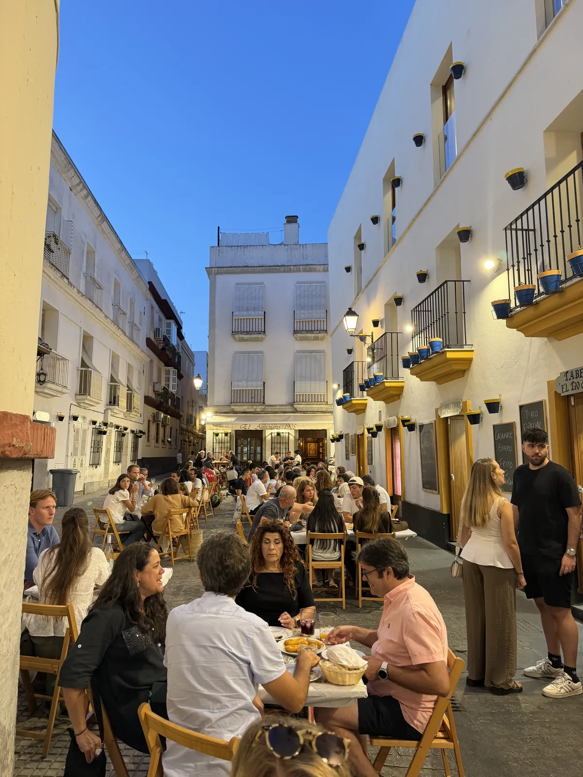 Volle Altstadtgasse in Cádiz am Abend: Menschen sitzen an Tischen vor weißen Häusern, darüber blauer Himmel.
