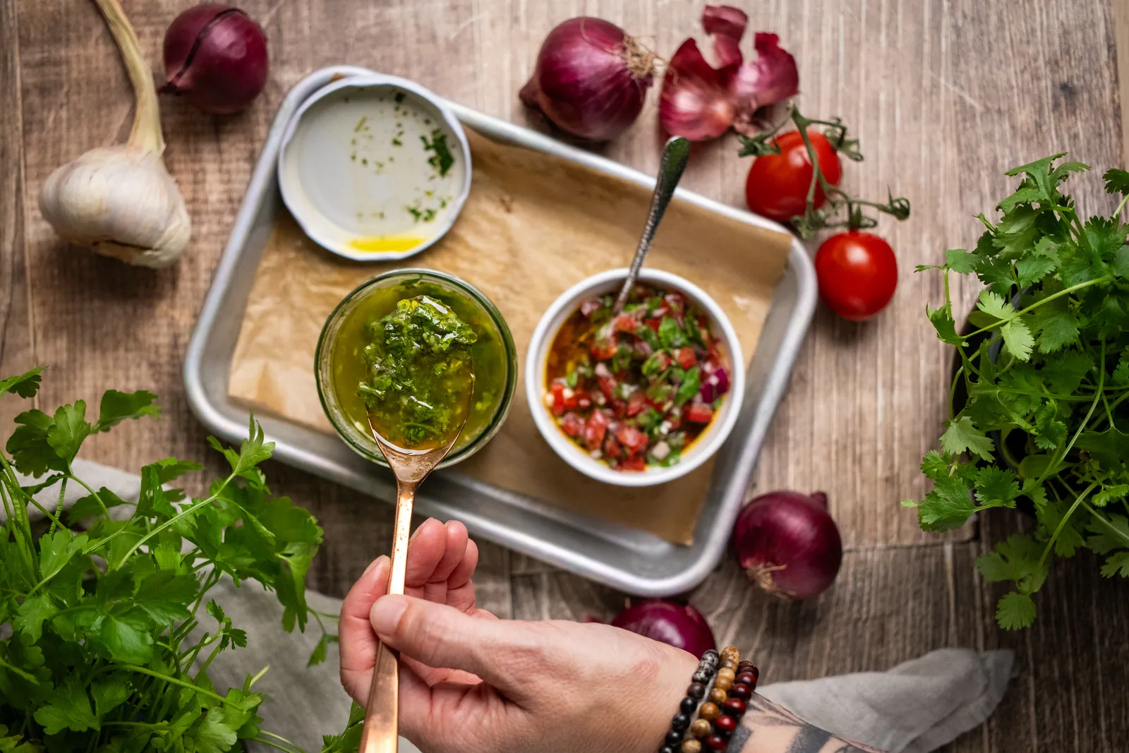 Chimichurri und Salsa Fresca in Schälchen auf einem Tablett, daneben Petersilie, Tomaten und rote Zwiebeln.