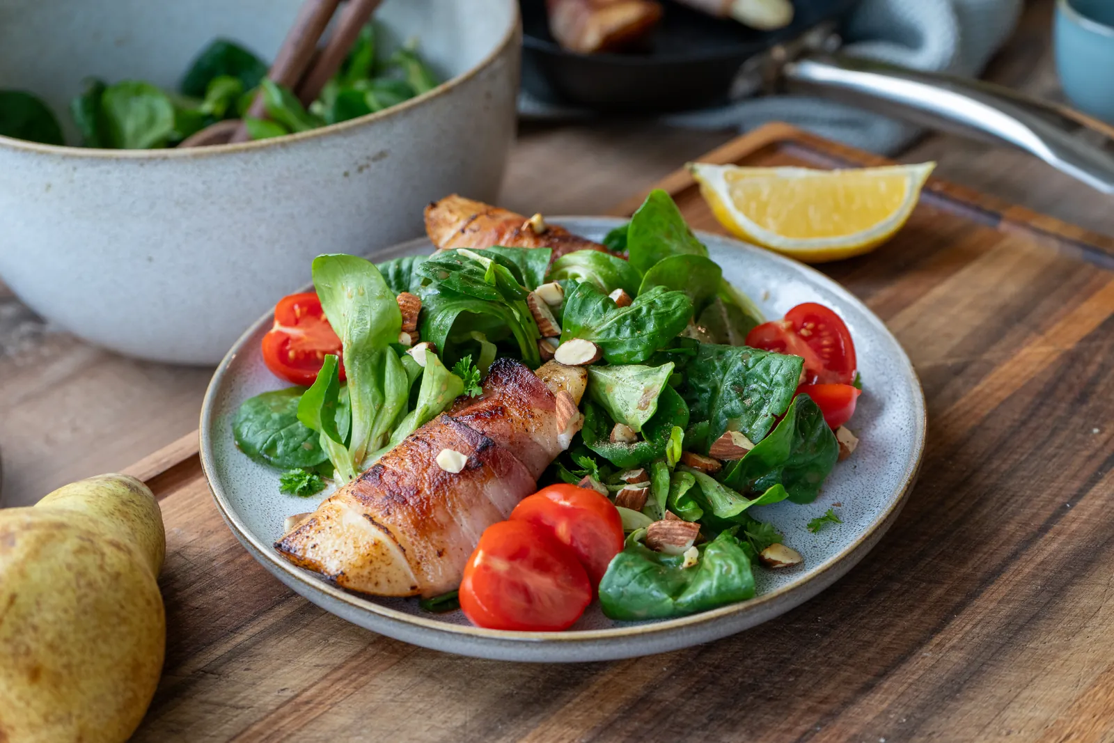 Feldsalat mit Birne im Speckmantel, Tomaten und Mandeln auf einem Teller, daneben eine Salatschüssel und eine ganze Birne auf einem Holzbrett.
