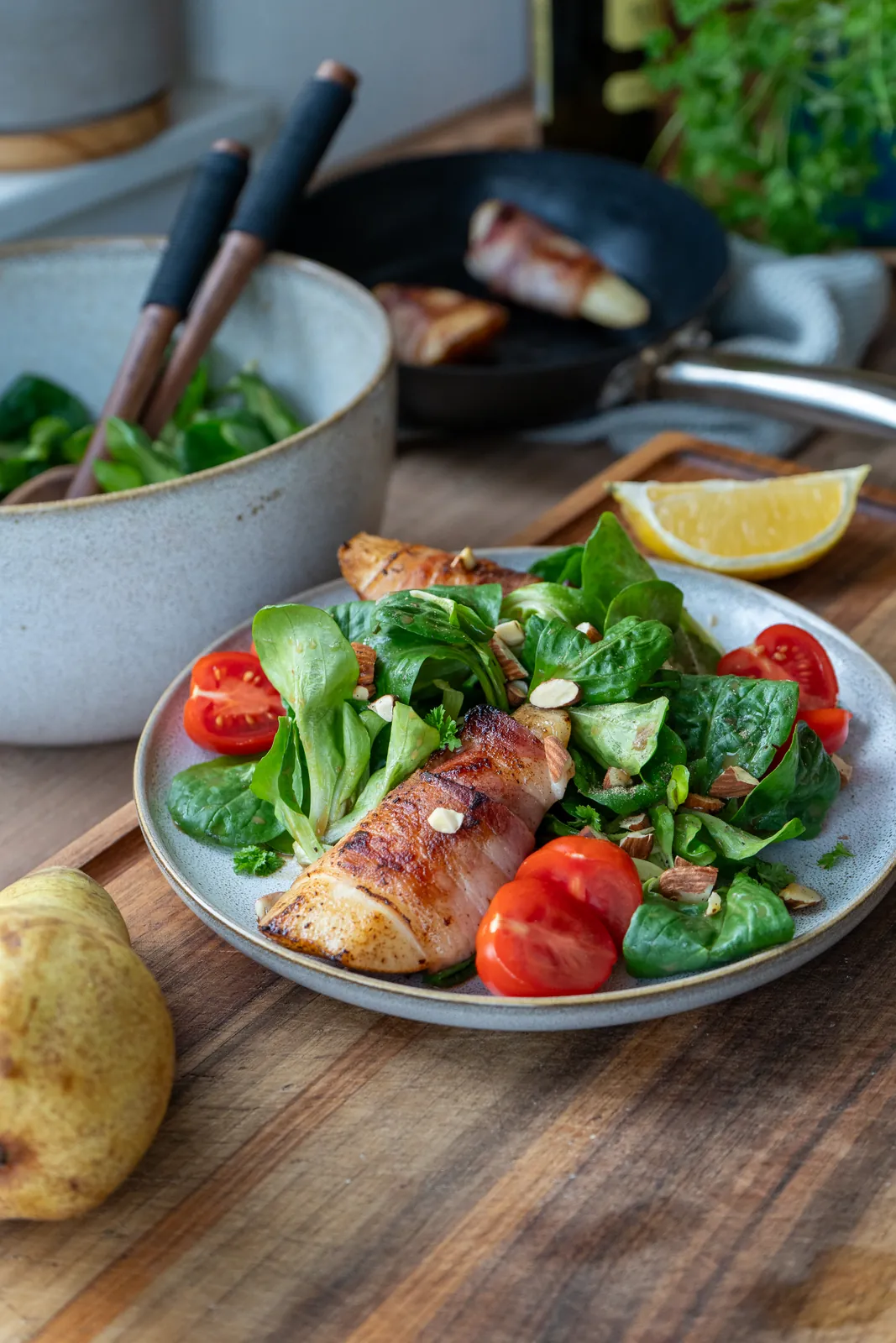 Feldsalat mit Speckbirne und Tomaten im Hochformat, dahinter Salatschüssel, Pfanne und Zitronenspalte.