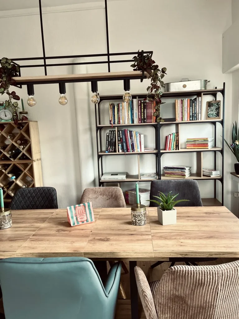 Lange Holztafel mit Stühlen unter einer Lampenschiene, dahinter ein Bücherregal