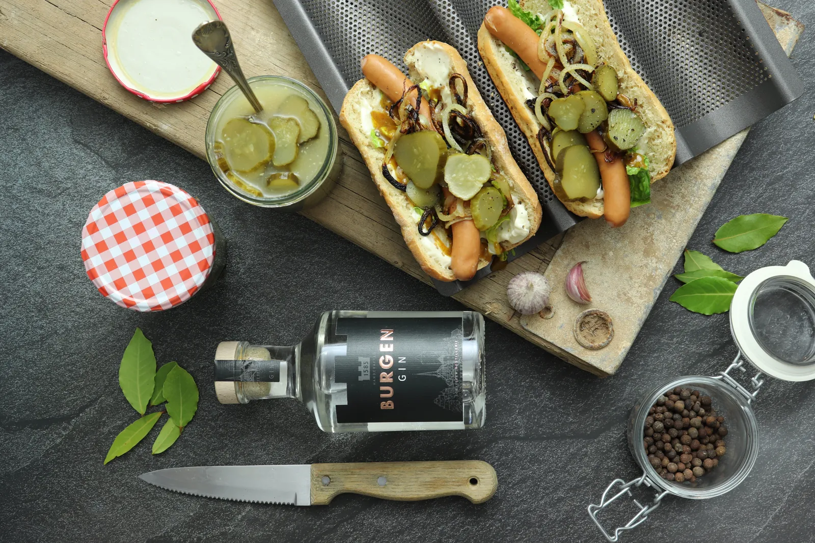Hot Dogs mit Gin-Gurken auf einem Holzbrett, daneben ein Glas Gurken, eine Flasche Gin und ein Messer.