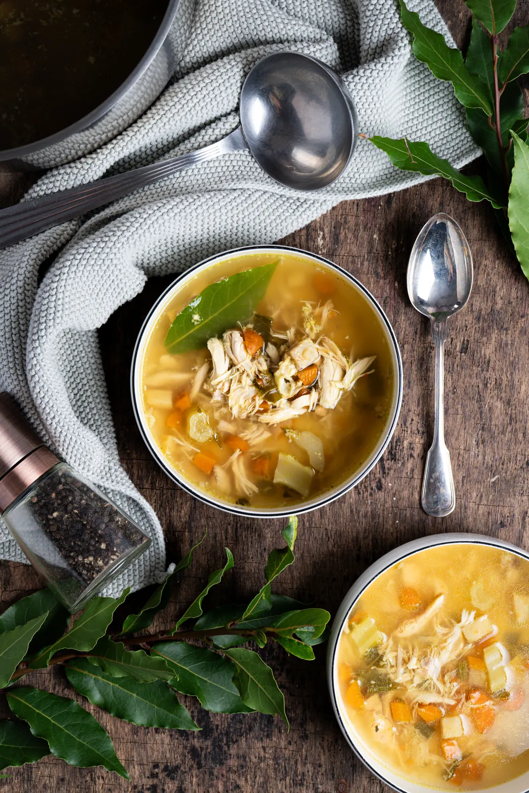 Hühnersuppe mit Gemüse und Lorbeerblatt in einer Schale von oben, daneben Kelle, Löffel und Lorbeerzweige auf dunklem Holz.