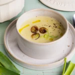 Schale mit heller Kartoffel-Lauch-Suppe, gebratenen Lauchringen, Olivenöl und Pfeffer auf einem Teller, daneben ein Löffel und Lauchstangen auf mintfarbenen Fliesen.