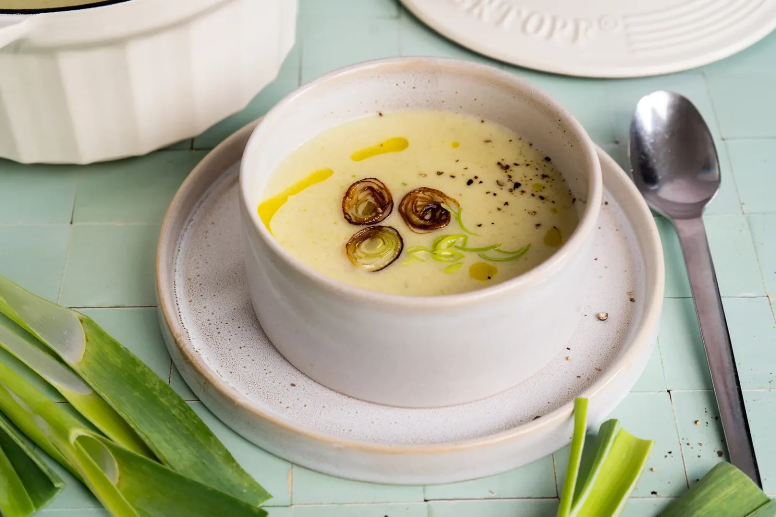 Schale mit heller Kartoffel-Lauch-Suppe, gebratenen Lauchringen, Olivenöl und Pfeffer auf einem Teller, daneben ein Löffel und Lauchstangen auf mintfarbenen Fliesen.