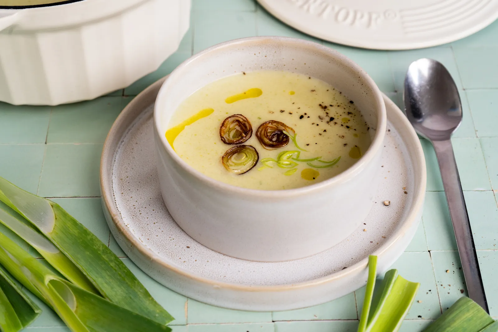 Schale mit heller Kartoffel-Lauch-Suppe, gebratenen Lauchringen, Olivenöl und Pfeffer auf einem Teller, daneben ein Löffel und Lauchstangen auf mintfarbenen Fliesen.