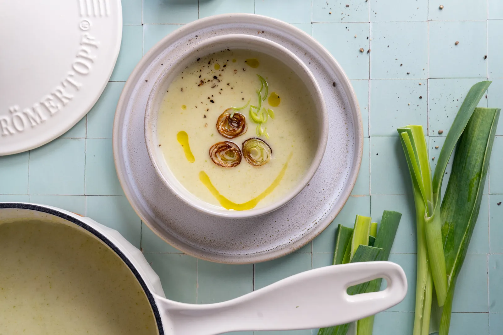 Kartoffel-Lauch-Suppe von oben in einer Schale, daneben ein weißer Gusseisentopf mit Suppe und frischer Lauch.