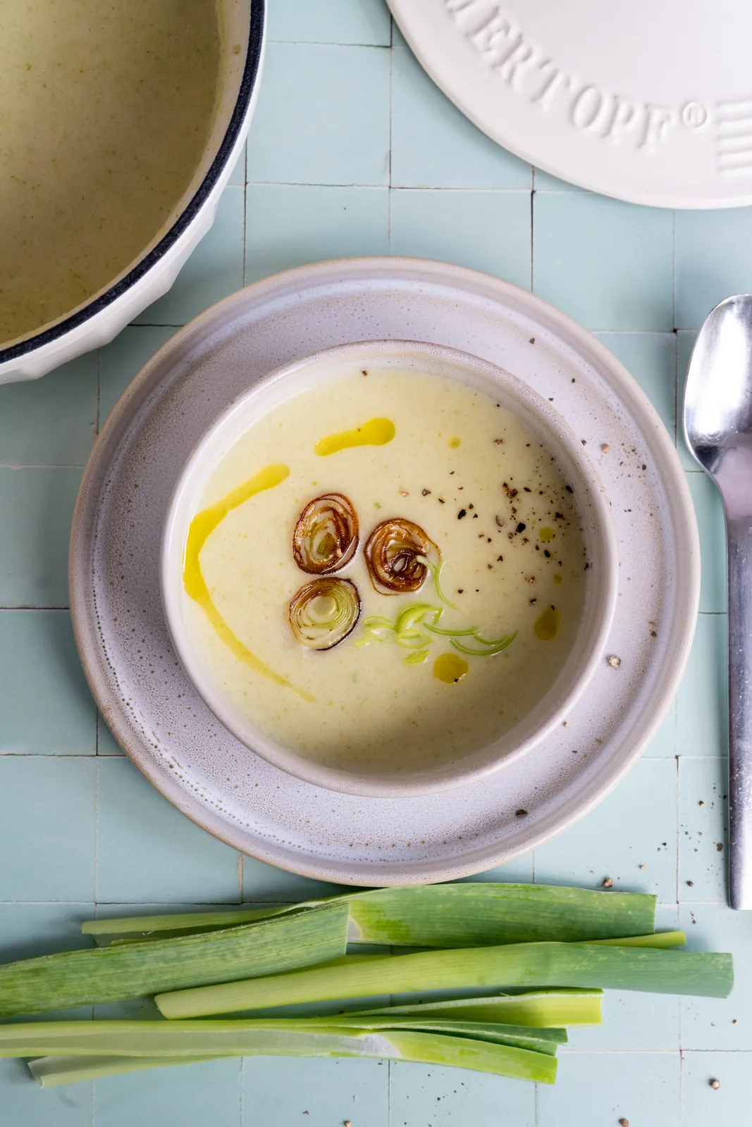 Kartoffel-Lauch-Suppe mit Lauchringen und Olivenöl im Hochformat, unten frischer Lauch, oben der Deckel eines Gusseisentopfs.
