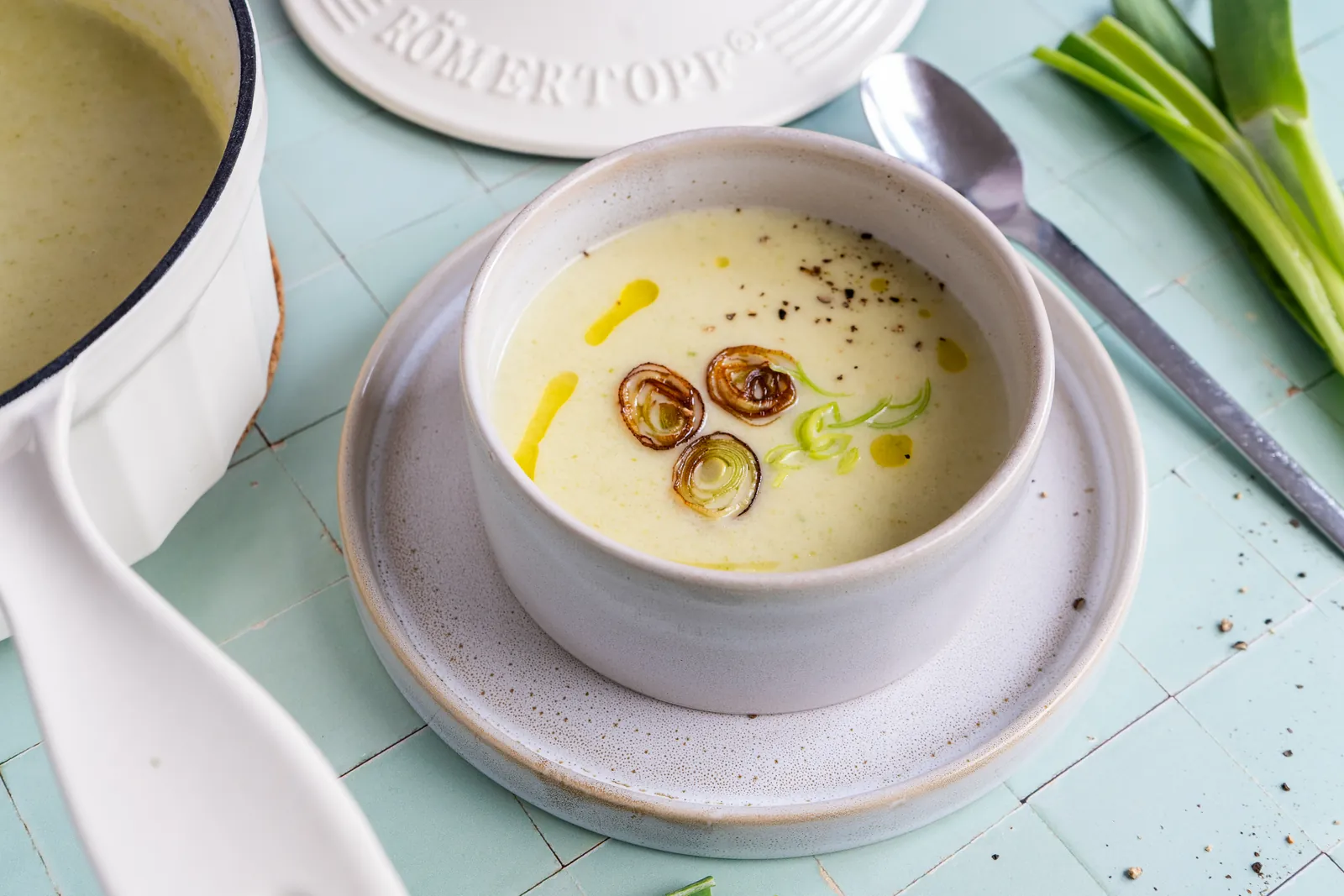 Schale Kartoffel-Lauch-Suppe mit Löffel auf einem Teller, im Hintergrund ein weißer gusseiserner Topf und Deckel.