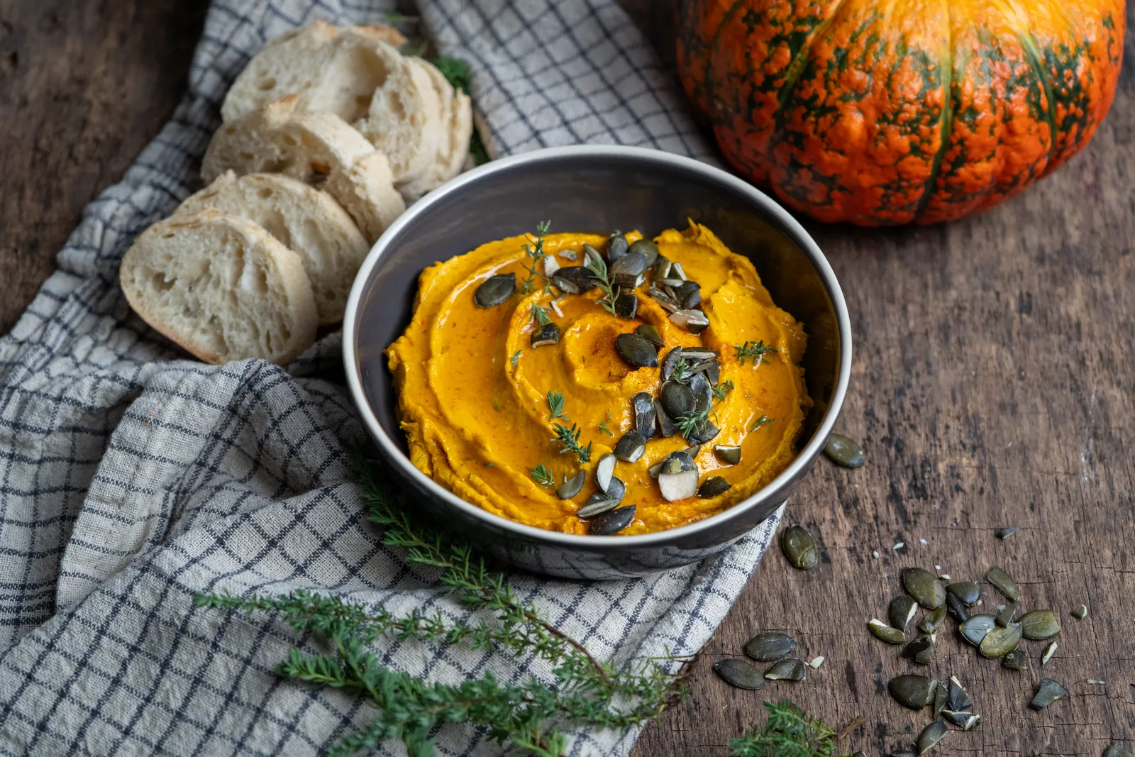 Kürbis-Dip mit Kürbiskernen und Thymian in einer Schale auf einem karierten Tuch, daneben Baguettescheiben und ein orangefarbener Kürbis.