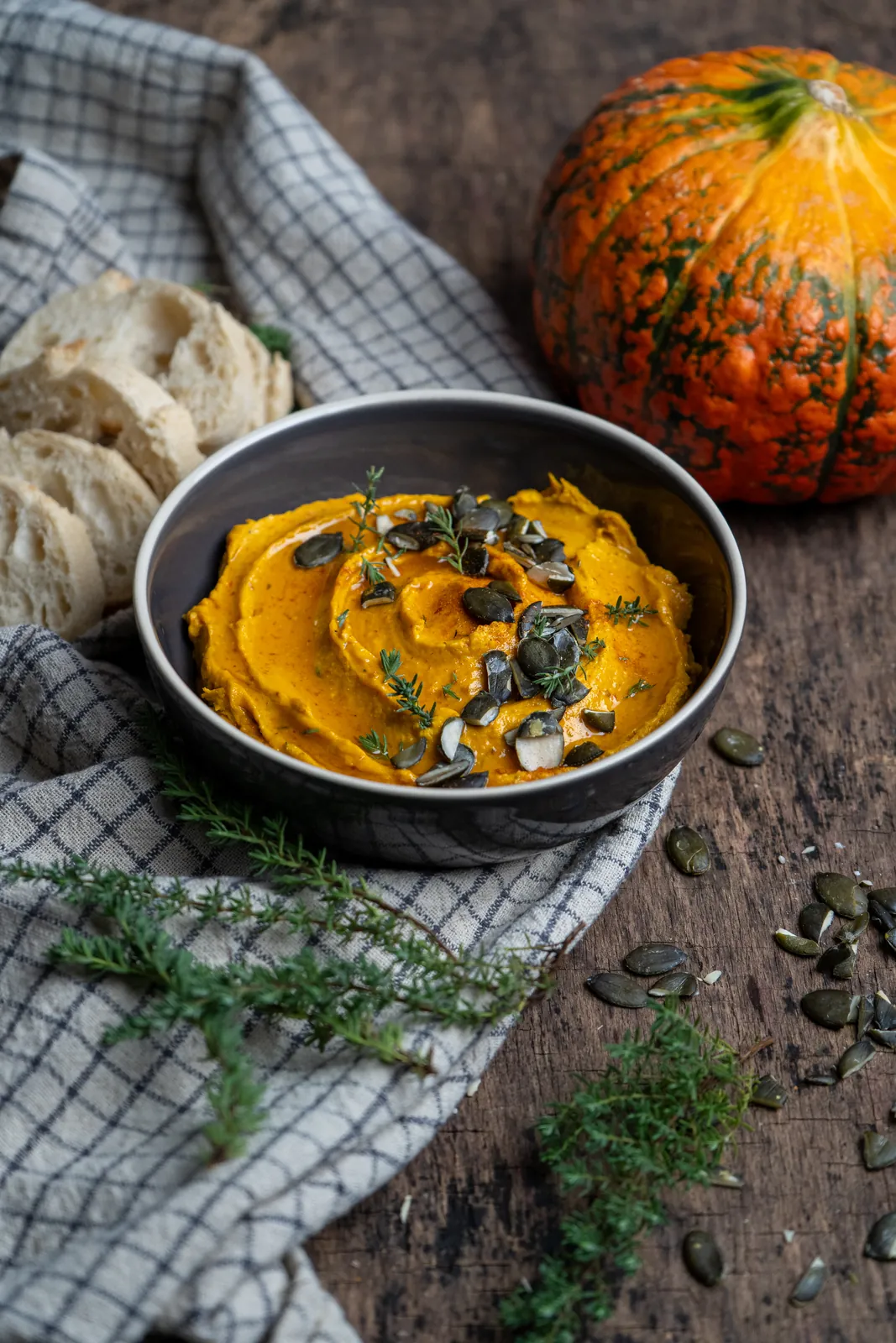 Schale mit orangefarbenem Kürbis-Dip, Kürbiskernen und Thymian auf dunklem Holz, Hochformat.
