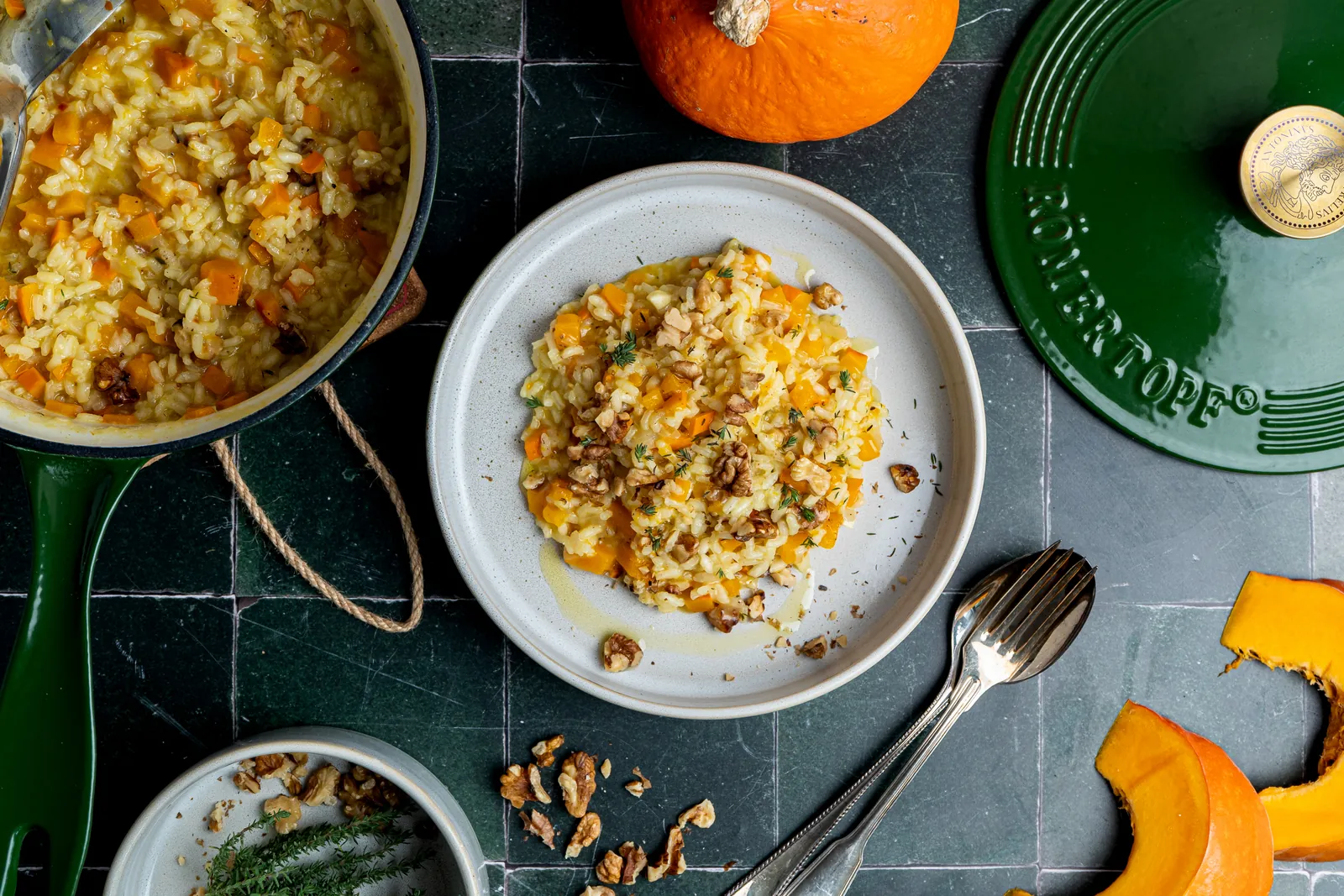 Kürbisrisotto mit Walnüssen auf einem weißen Teller, daneben ein grüner gusseiserner Topf mit Risotto, Hokkaido-Kürbis und Thymian, von oben fotografiert.