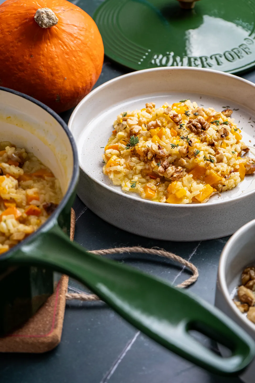 Teller mit Kürbisrisotto und Walnüssen, im Vordergrund der Griff einer grünen gusseisernen Stielkasserolle, daneben ein Hokkaido.