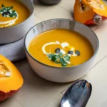 Zwei Schalen orangefarbene Kürbissuppe mit Sahne, Kürbiskernen und Petersilie, daneben Hokkaido-Stücke und Löffel auf hellen Fliesen.