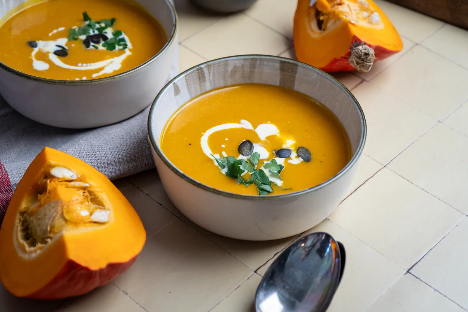 Zwei Schalen orangefarbene Kürbissuppe mit Sahne, Kürbiskernen und Petersilie, daneben Hokkaido-Stücke und Löffel auf hellen Fliesen.