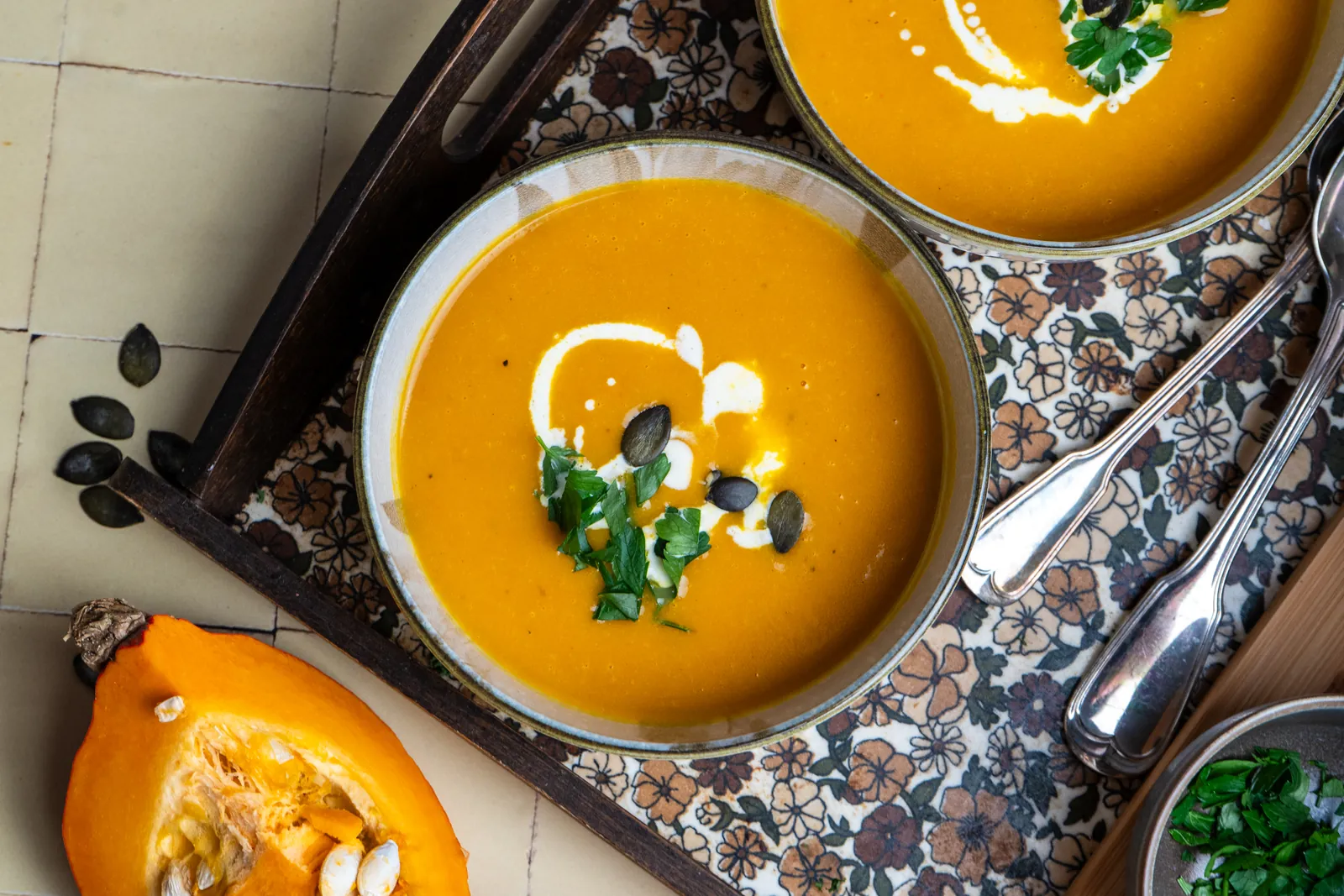 Schale Kürbissuppe mit Sahneschliere und Petersilie auf einem Tablett mit Blumenmuster, daneben Hokkaido-Spalten.