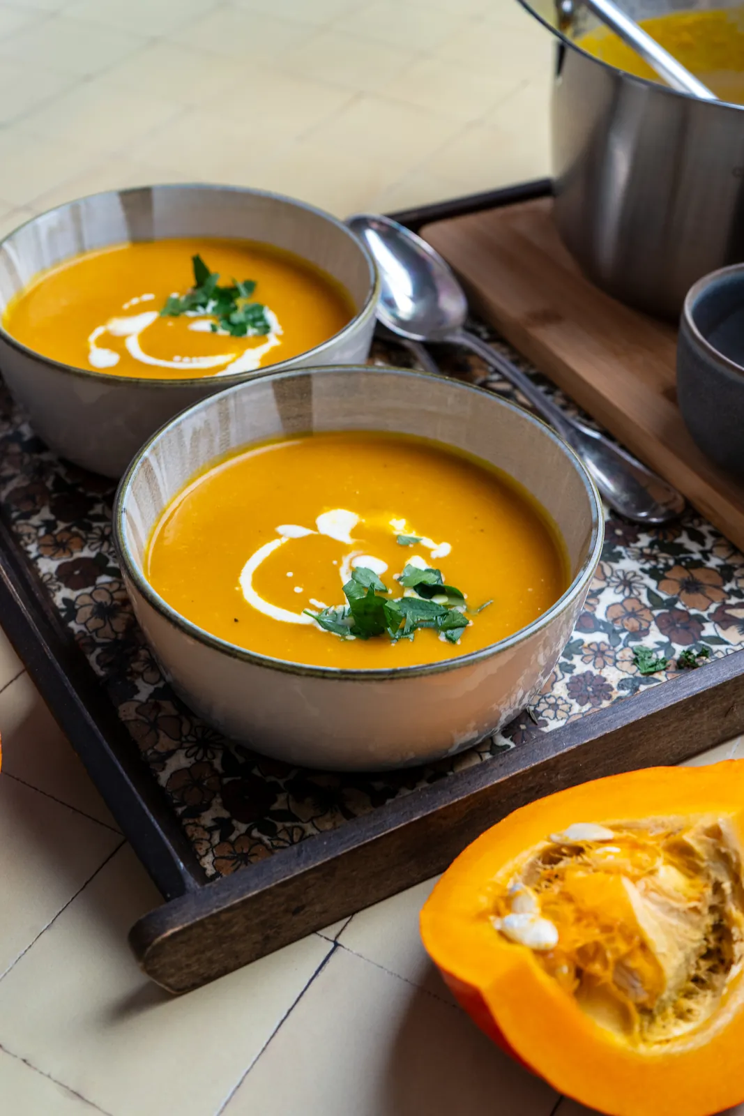 Kürbissuppe in Schalen auf einem Tablett, daneben ein Topf, Löffel und ein Stück Hokkaido.