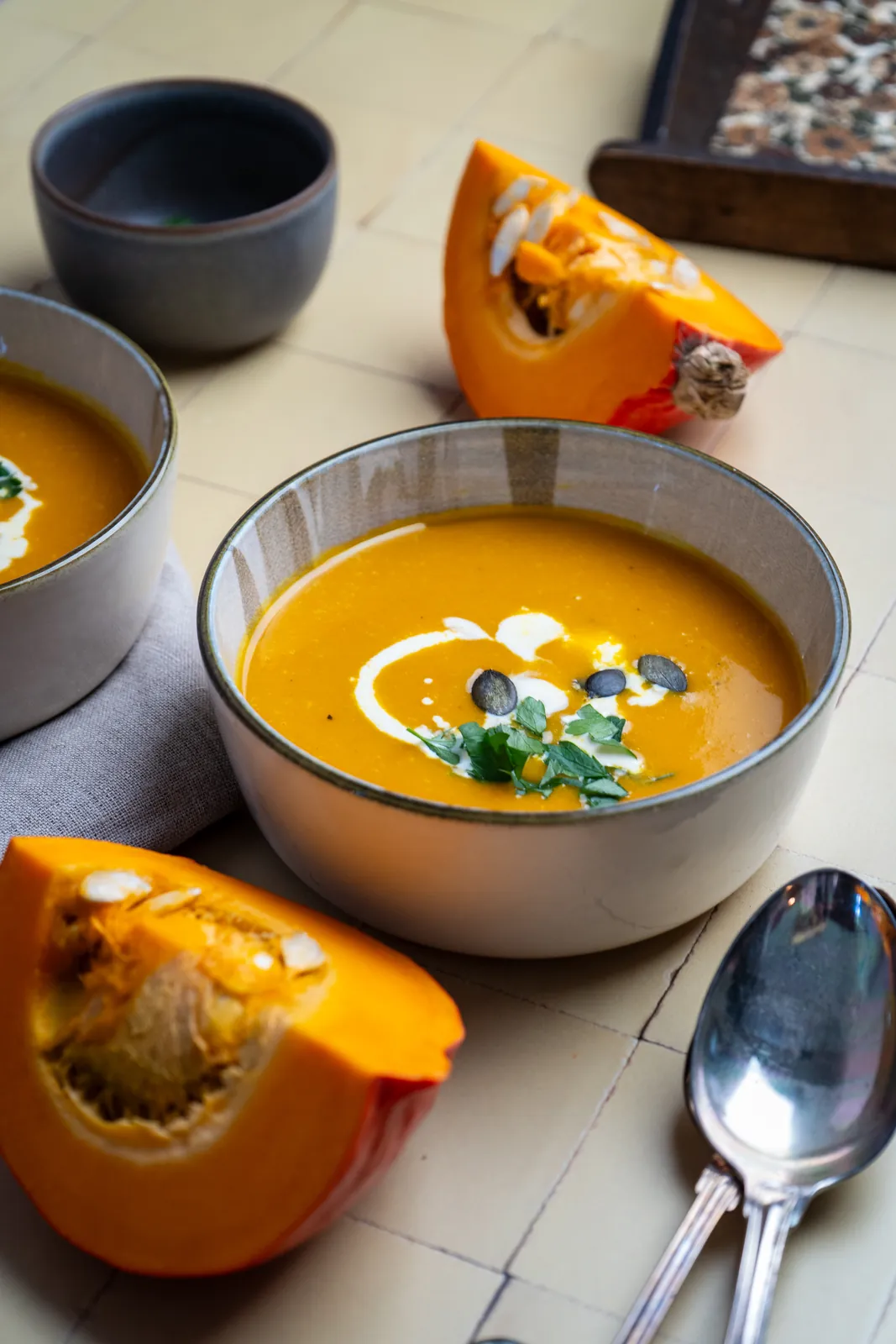 Schale mit Kürbissuppe, Sahne und Petersilie von oben, daneben Hokkaido-Stücke und zwei Löffel.
