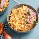 Schale mit Obatzda, garniert mit roten Zwiebelringen und Petersilie, daneben eine Brezel und Schalotten auf hellblauem Untergrund.