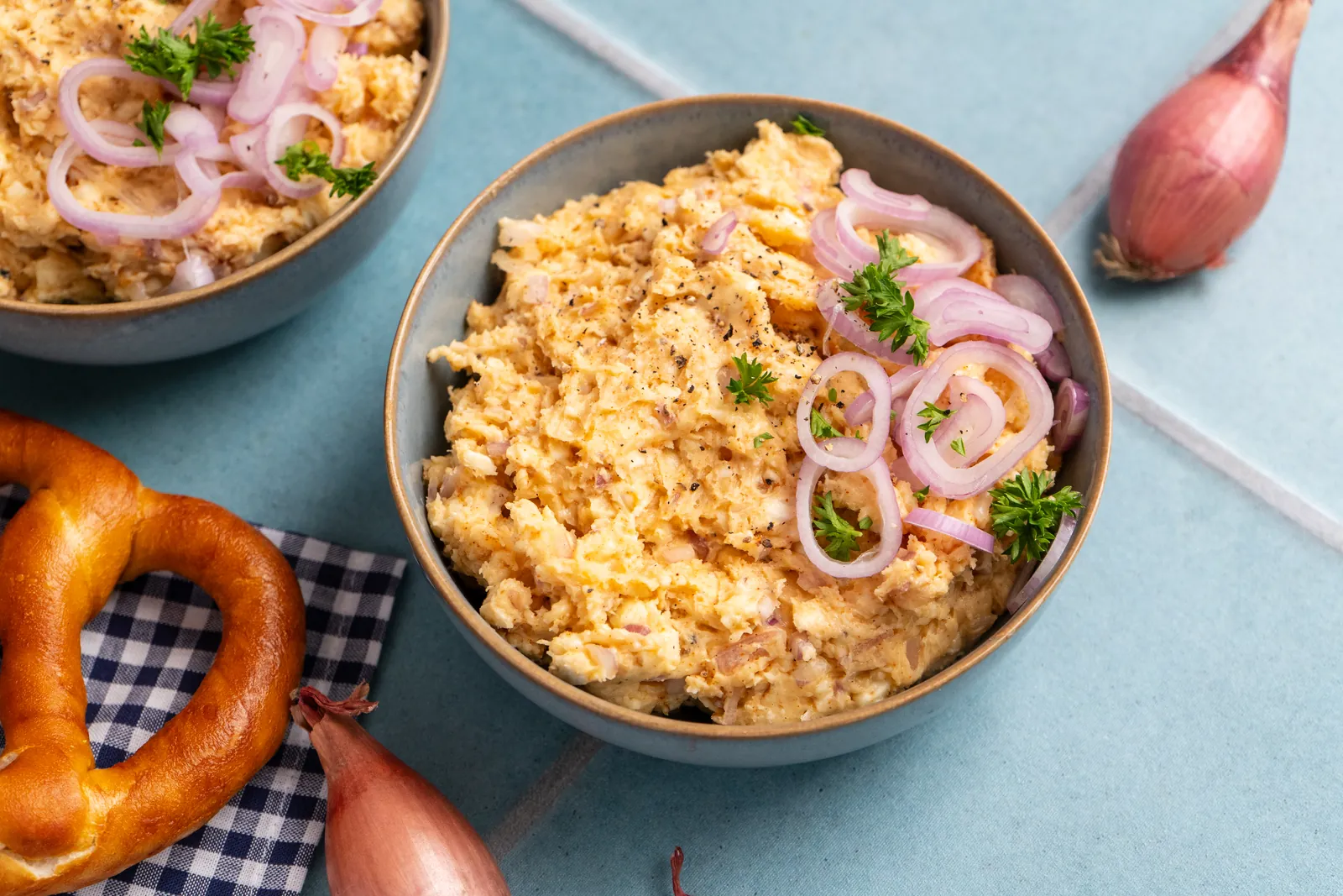Schale mit Obatzda, garniert mit roten Zwiebelringen und Petersilie, daneben eine Brezel und Schalotten auf hellblauem Untergrund.