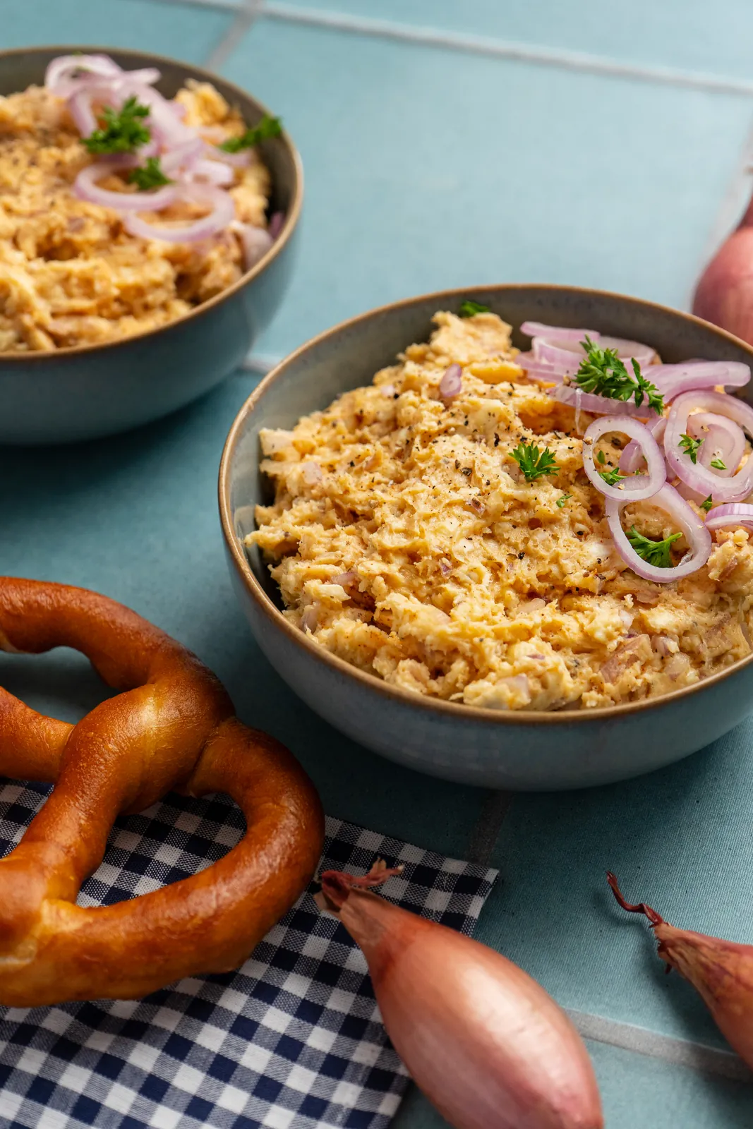 Obatzda in einer Schale mit Zwiebelringen und Petersilie, daneben Brezel, Schalotten und ein karierter Stoff.