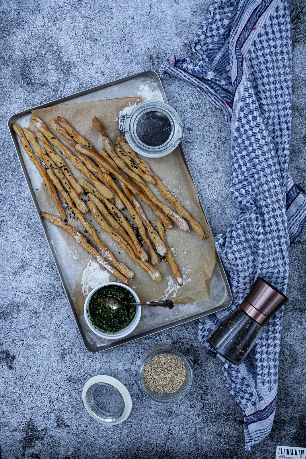 Selbstgemachte Thymian-Grissini auf einem Backblech auf grauem Untergrund, daneben Pesto und eine Pfeffermühle.