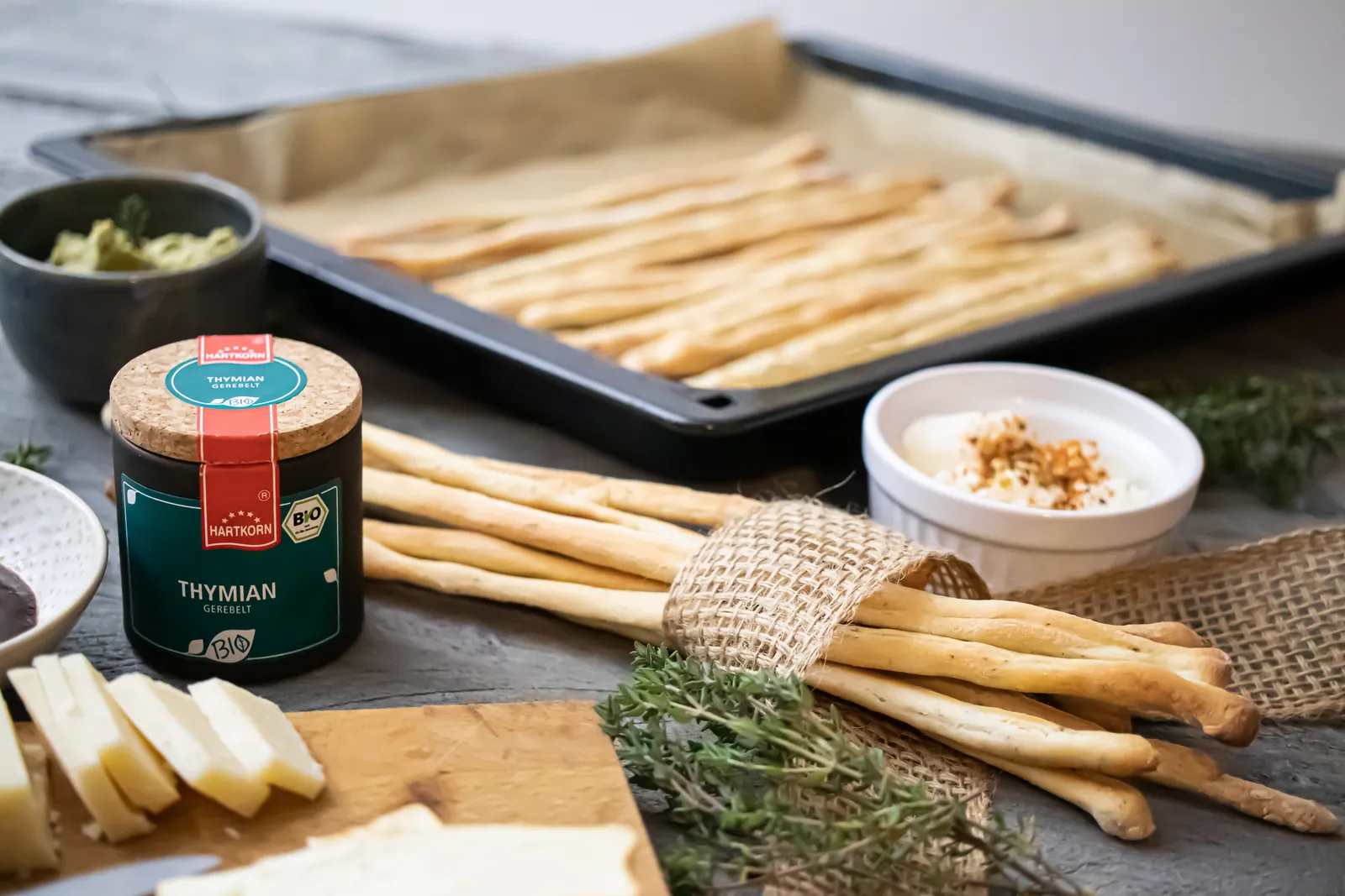 Thymian-Grissini auf einem Backblech und gebündelt auf einem Brett, davor Käse und ein Dip.