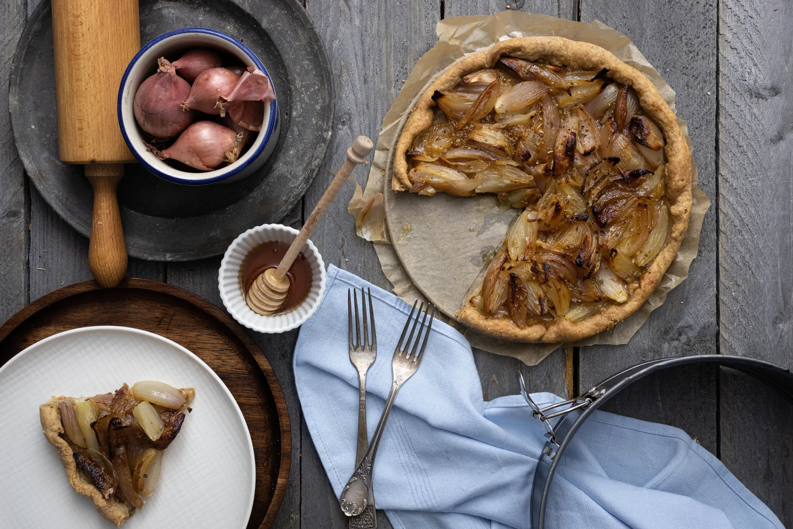 Zwiebelkuchen mit Schalotten, ein Stück auf einem Teller, daneben Teigrolle und Schalotten, von oben.