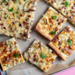 Zwiebelkuchen vom Blech in Stücke geschnitten auf Backpapier, mit Speckwürfeln und Petersilie, von oben fotografiert.
